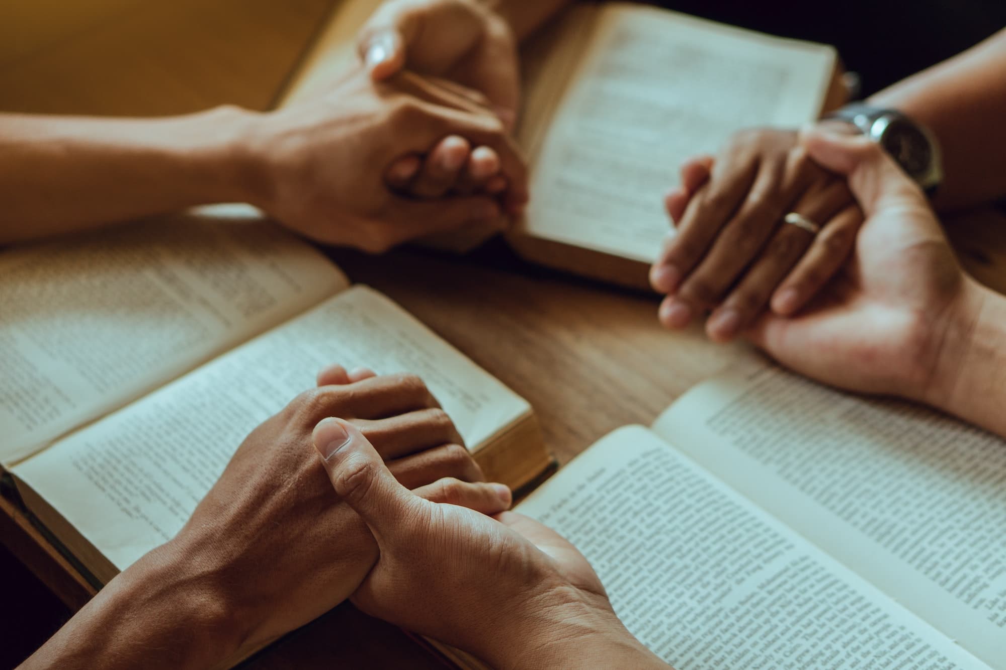 Hands folded in prayer over open Bibles around a wooden table